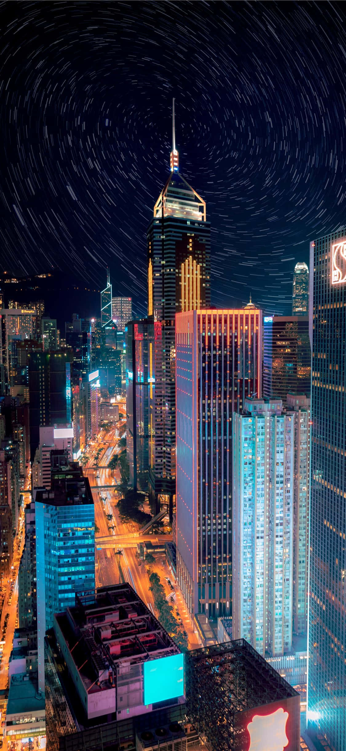 探索永不眠的紐約城 — iPhone XS 紐約之旅 壁紙 — 一個令人眼花繚亂的夜景城市景觀，高聳的摩天大樓在黑暗天空中旋轉的星軌下閃耀。