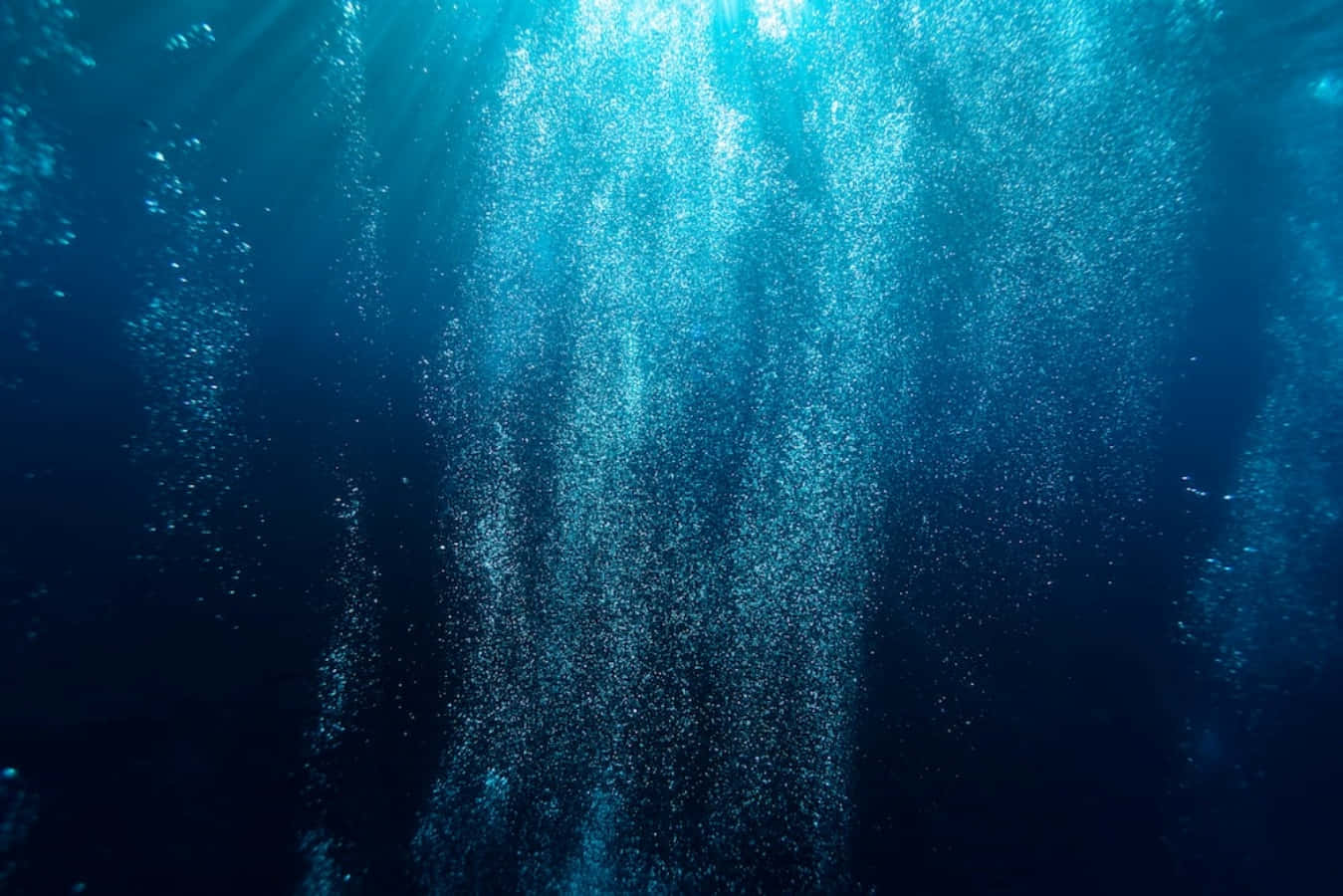 探索深海 壁紙 — 陽光光束穿透深藍色海水，照亮向上升起的旋渦氣泡流。