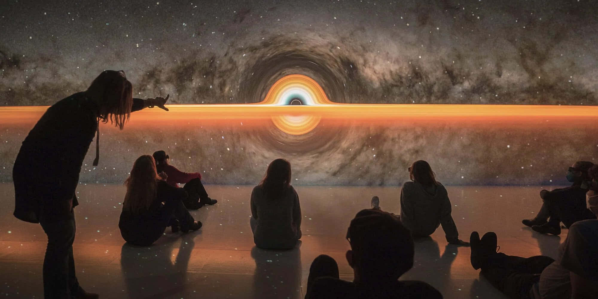 探索深邃的深處 壁紙 — 剪影人物坐著並指向一個在星空宇宙展覽中巨大發光的黑洞投影。