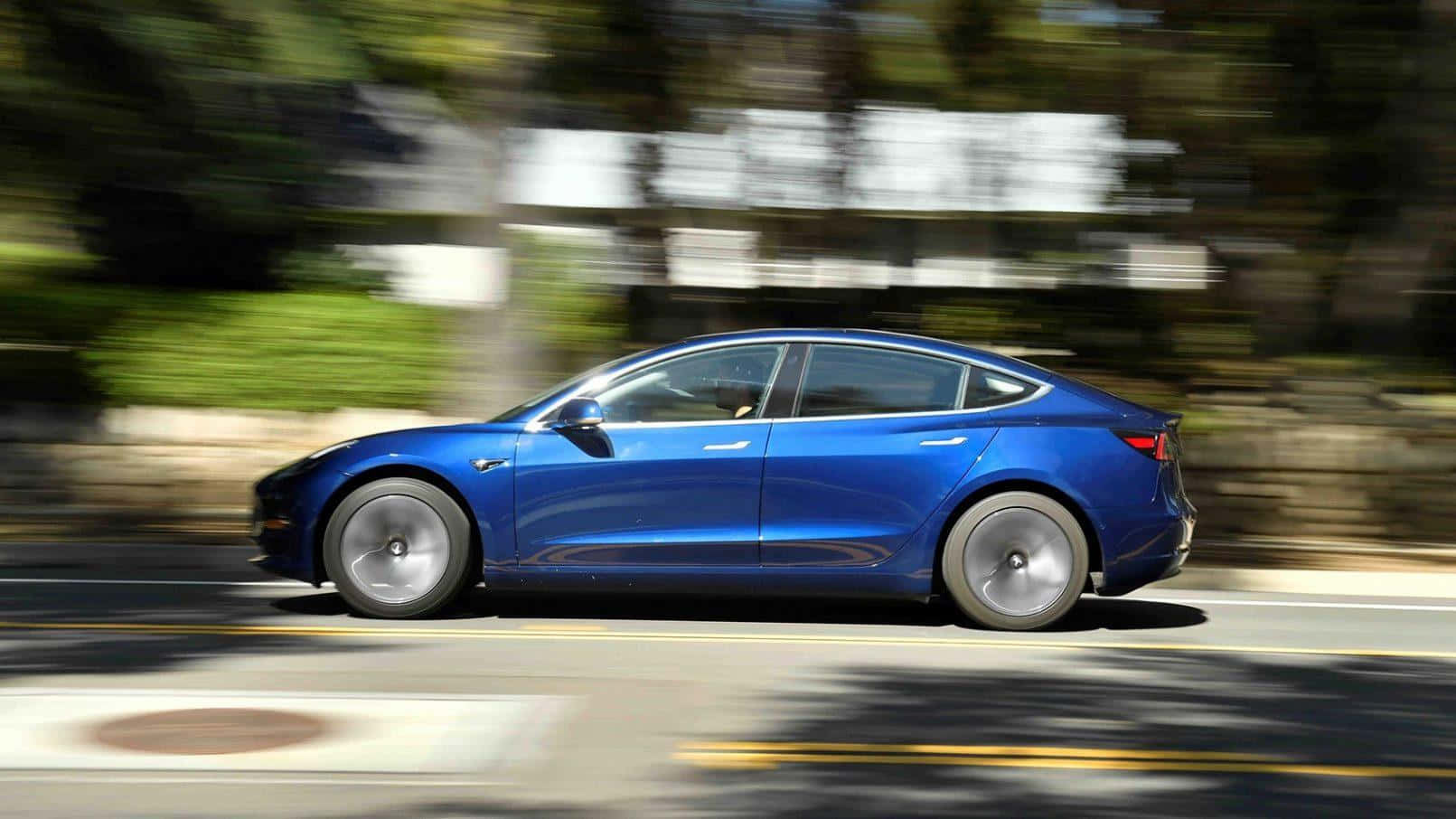 探索特斯拉的創新之作Model 3 壁紙 — 一輛充滿活力的藍色 Tesla Model 3 在陽光明媚的道路上行駛，模糊的綠色植物飛馳而過。