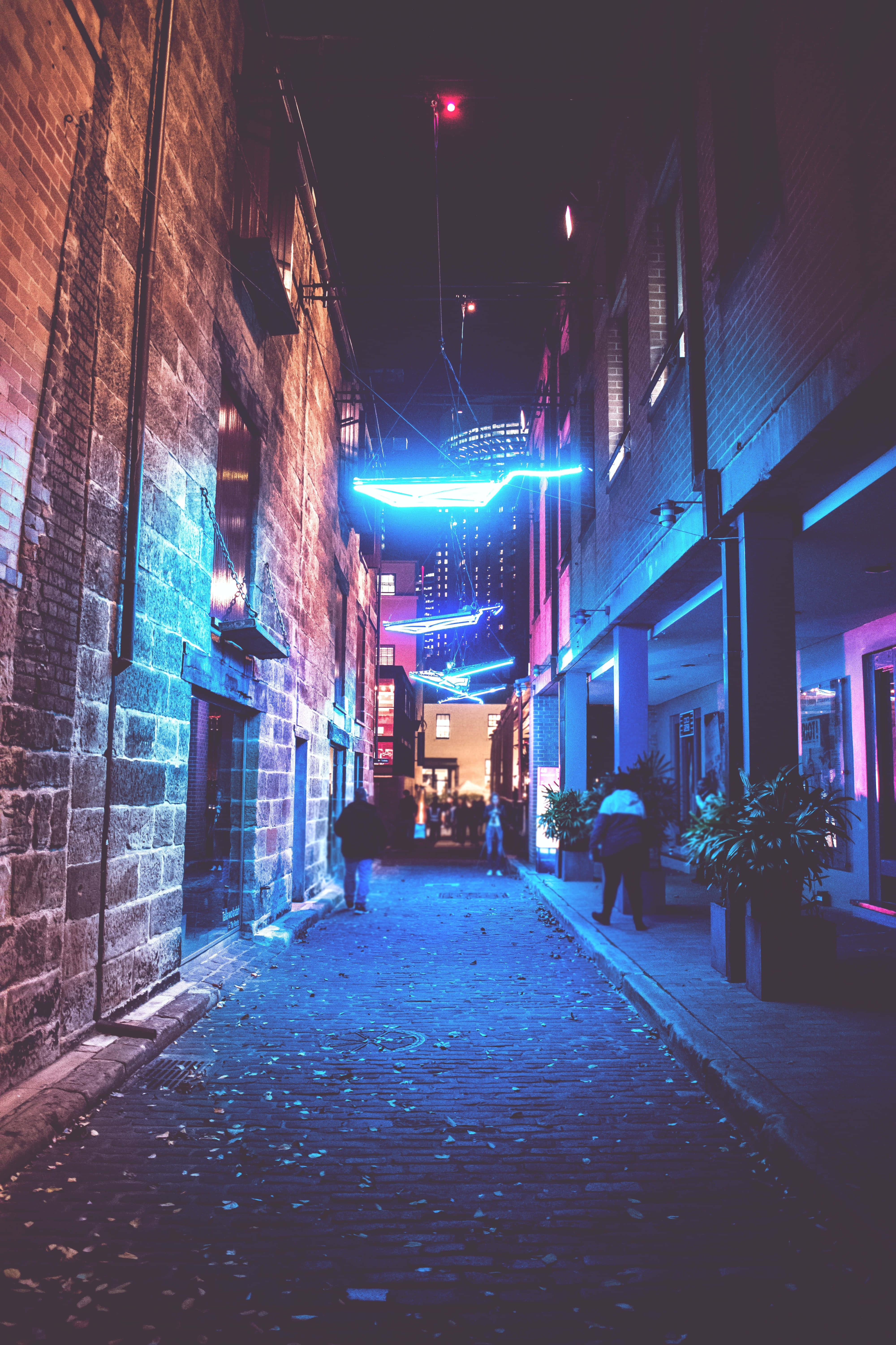「探索與體驗霓虹城市的未來感景觀」 壁紙 — 夜晚一條狹窄的磚砌小巷沐浴在充滿活力的藍色霓虹燈下，人們在發光的招牌下行走。