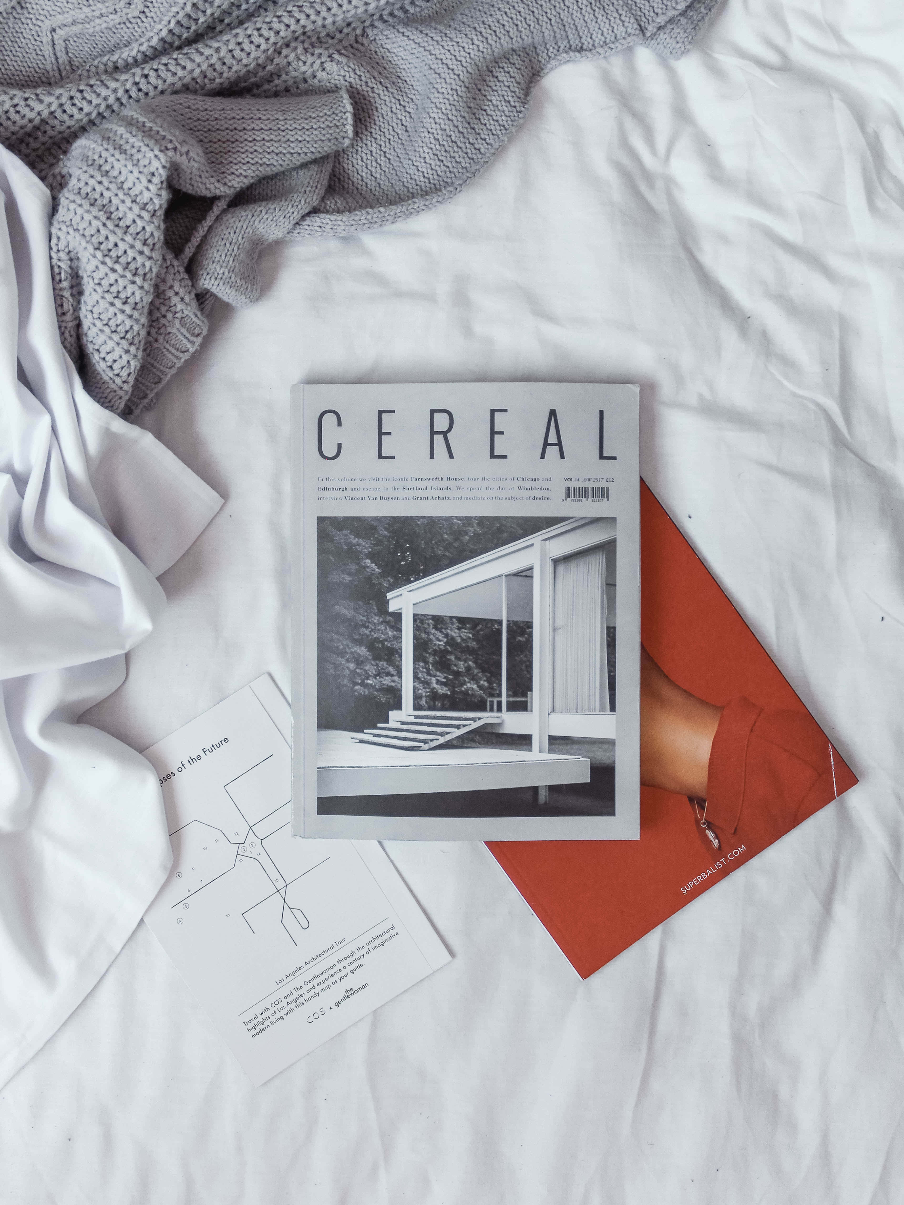探索雜誌，保持資訊靈通 壁紙 — 床上CEREAL雜誌、白色亞麻布、灰色針織品和橙色信封的極簡平鋪佈置。