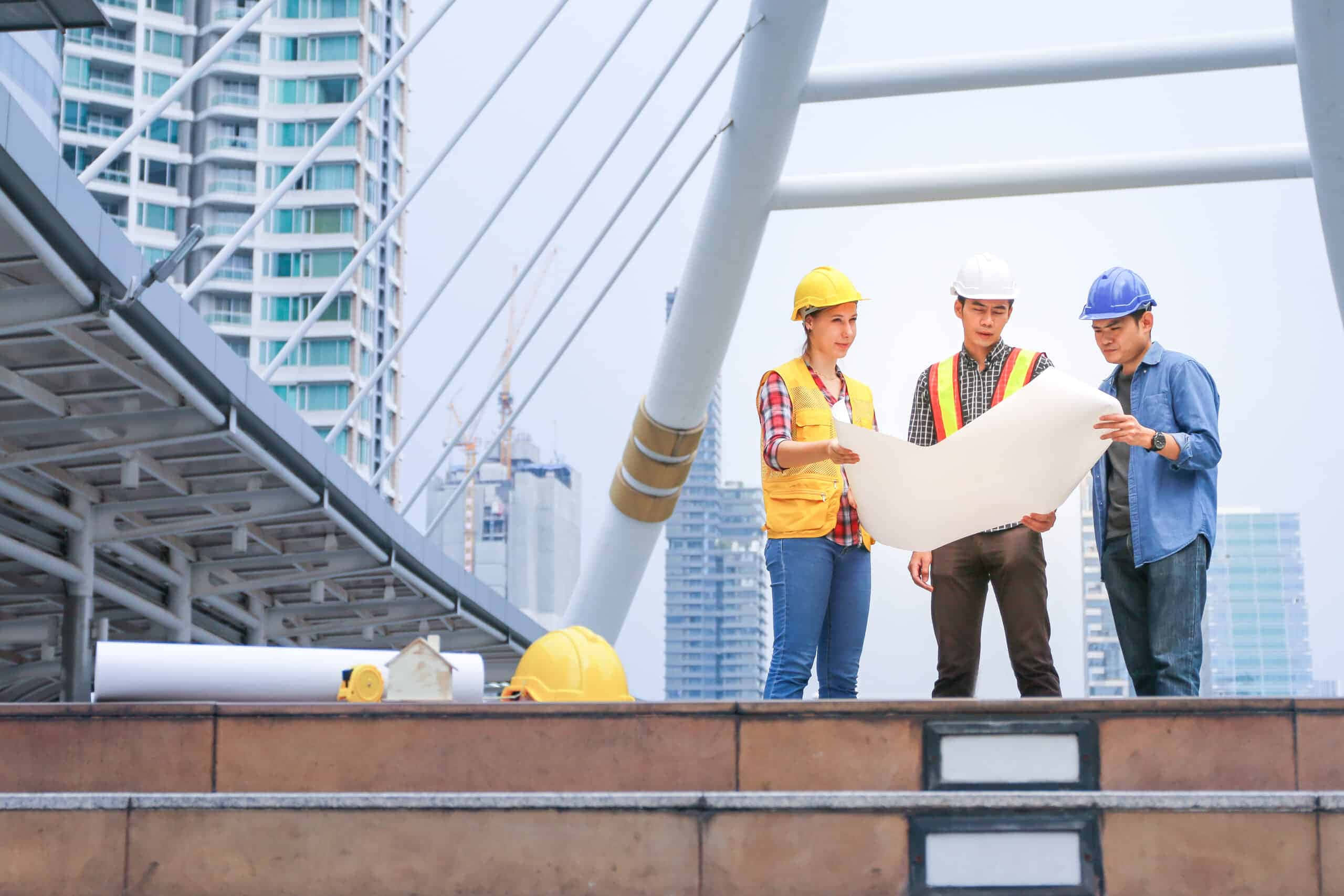 推動工程學的未來 壁紙 — 三名戴著安全帽的建築工人在一座現代橋樑上審閱藍圖，背景是高樓大廈。