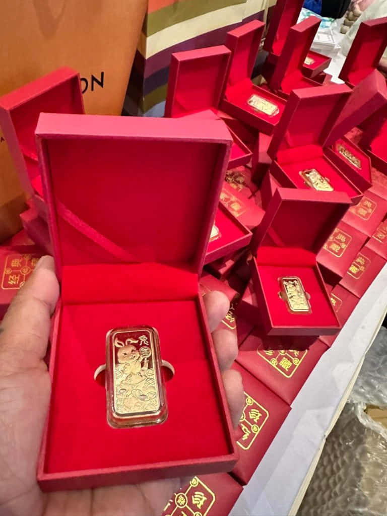 提升您的金條投資 壁紙 — 一隻手在商店中眾多相同的盒子中，拿著一個紅色盒子裡帶有兔子設計的金條。
