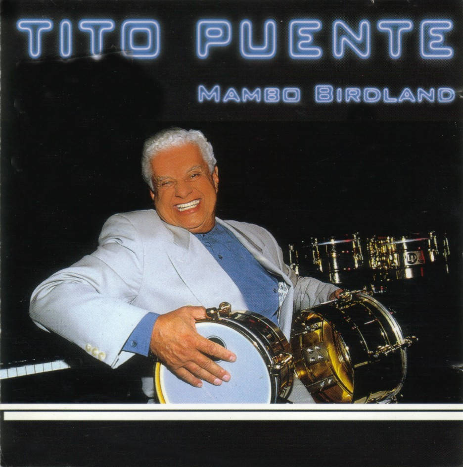 Tito Puente 在藍色霓虹文字的舞台上微笑著拿著鼓。