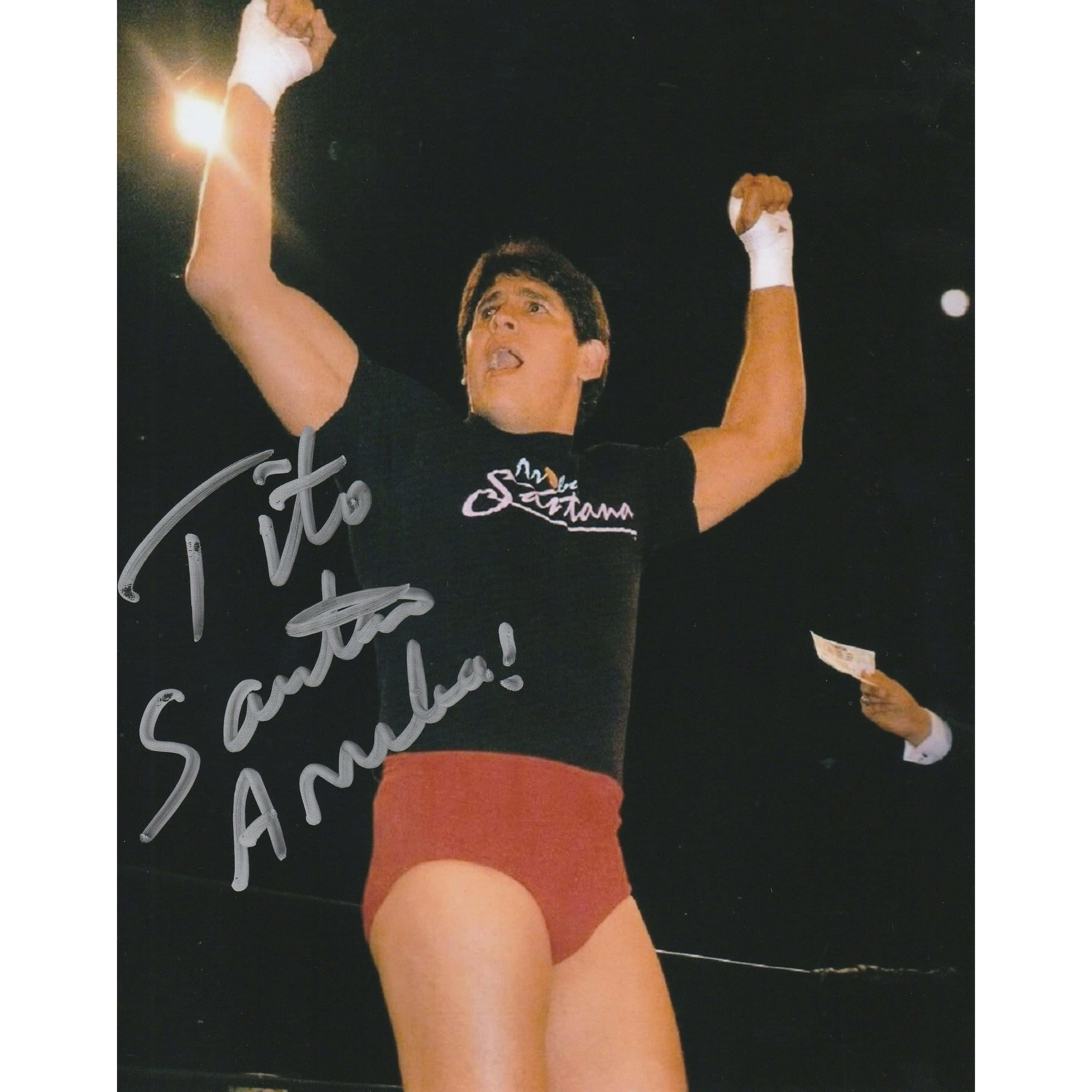 提托·桑塔納簽名勝利賽事照片 壁紙 — 摔角手 Tito Santana 在明亮的舞台燈光下，身穿黑色襯衫和紅色短褲，勝利地高舉雙拳。