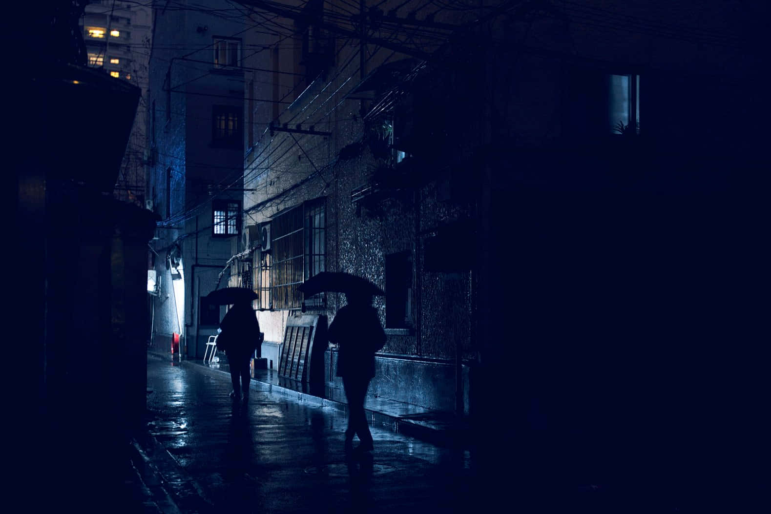 「揭開宇宙的祕密」 壁紙 — 兩個撐著雨傘的剪影人物在夜晚昏暗的藍色街燈下沿著濕滑的雨巷行走。
