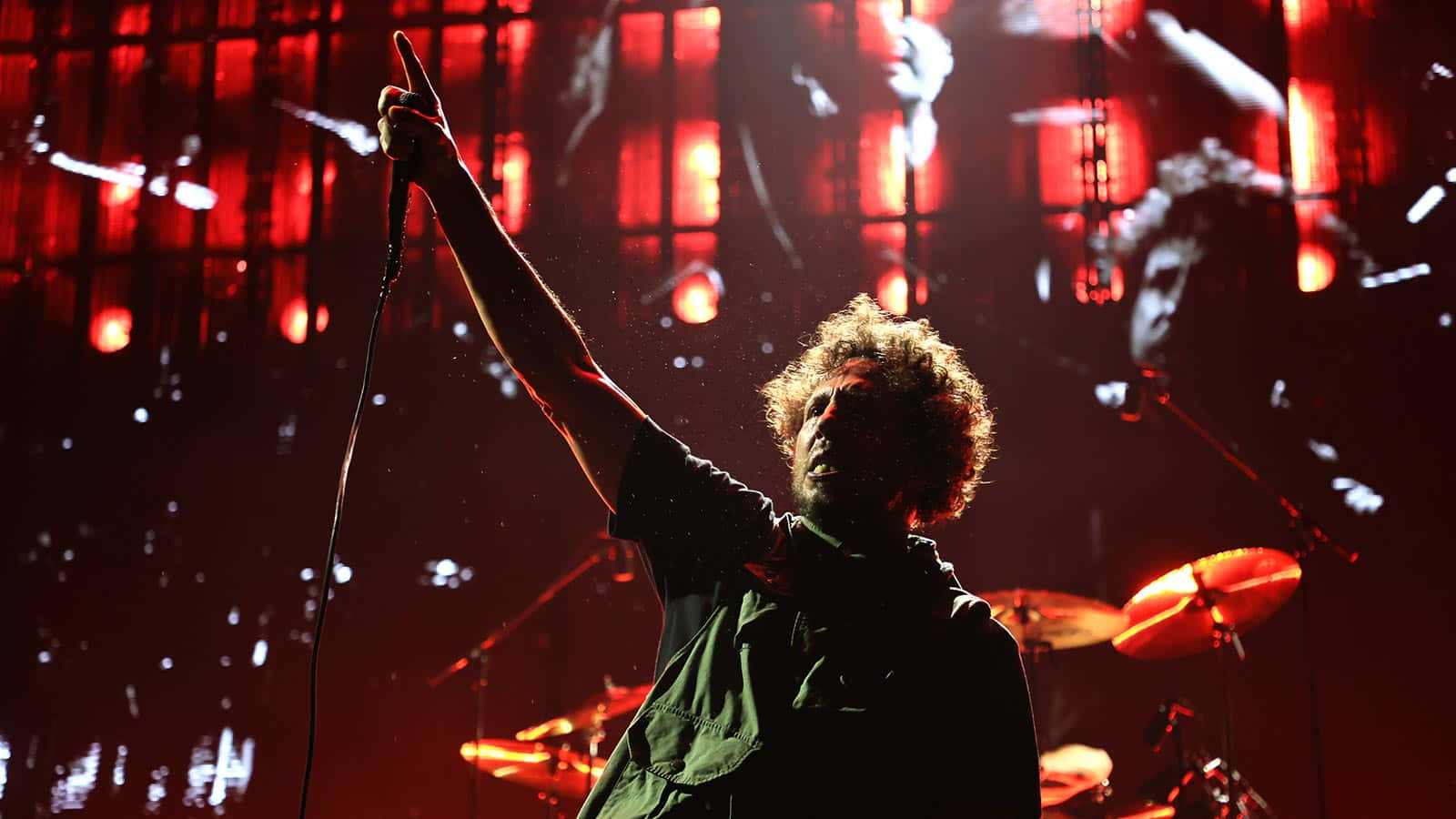 搖滾音樂會能量：Zack De La Rocha 壁紙 — 一位捲髮音樂家在強烈紅燈下於舞台上熱情演出，身後可見一套鼓。