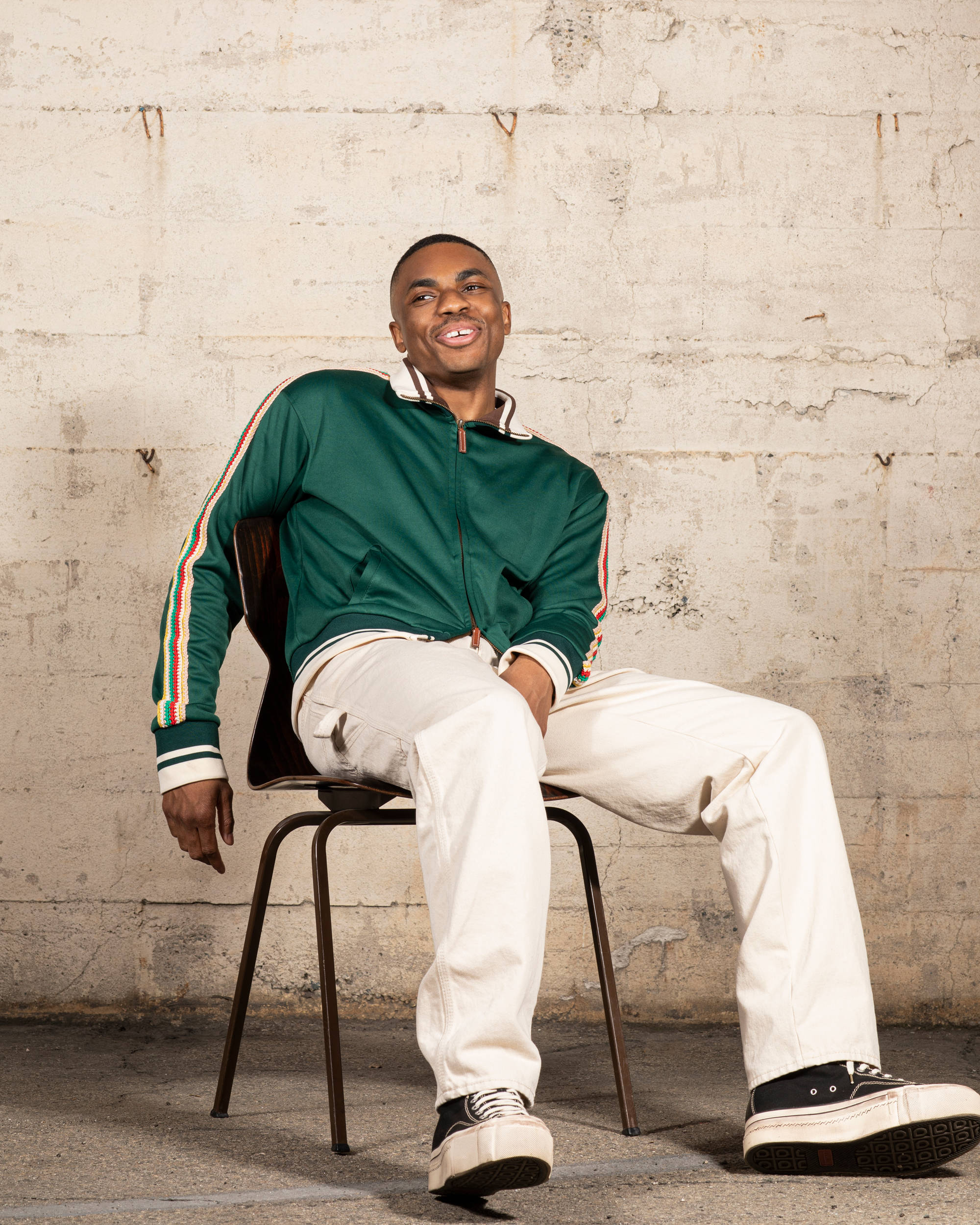 搞怪饒舌歌手 Vince Staples 壁紙 — 一位微笑的年輕男子穿著綠色運動外套和奶油色褲子，隨意地坐在金屬椅子上，背景是風化的混凝土牆。