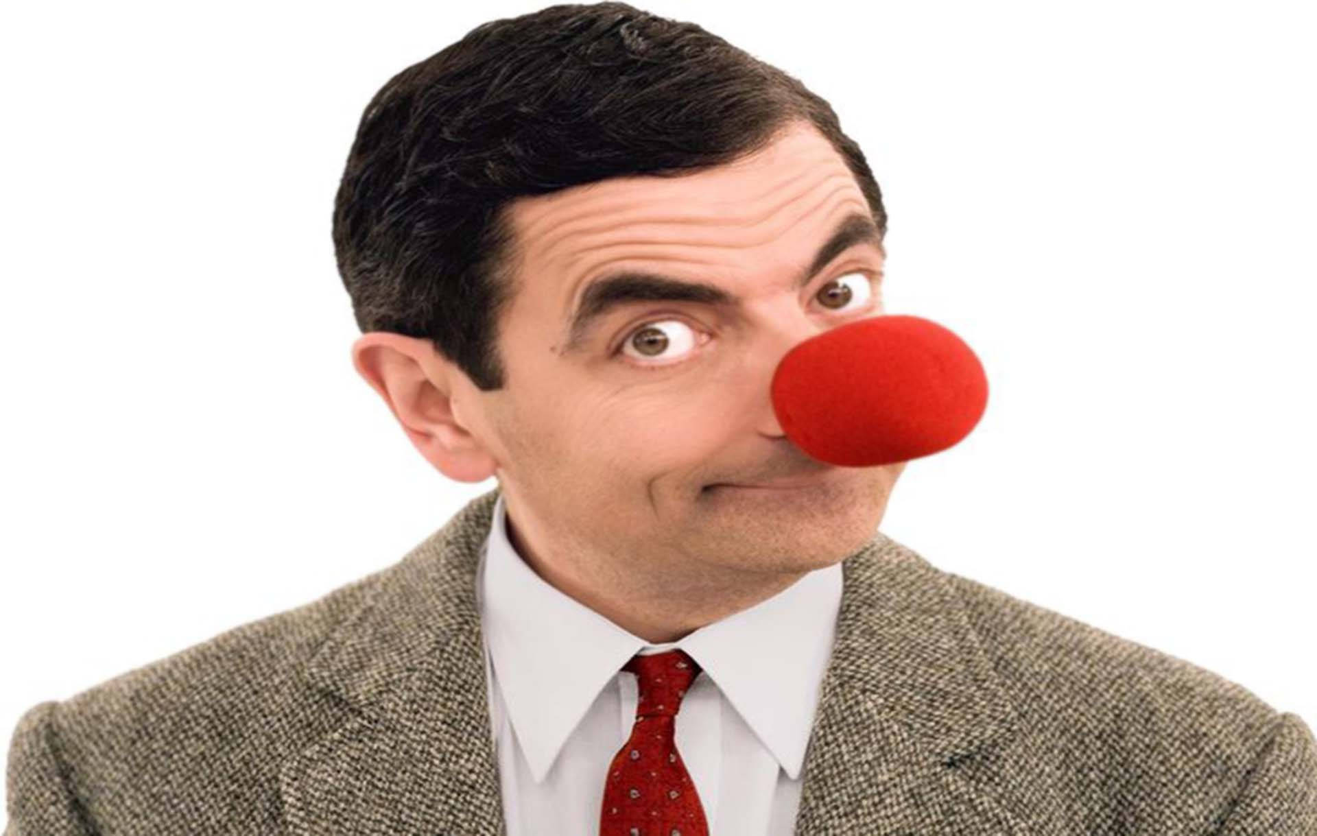 搞笑Mr. Bean戴紅鼻 壁紙 — Mr. Bean 戴著紅色小丑鼻子，穿著正式西裝，在純白背景前瞪大眼睛凝視。