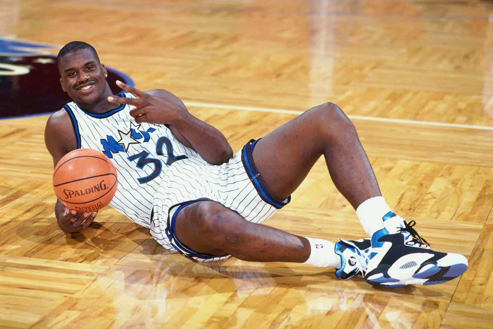 搞笑NBA和平手勢照 壁紙 — 一名微笑的籃球運動員身穿白色條紋球衣，號碼32，躺在木地板上，手持Spalding籃球並做出和平手勢。