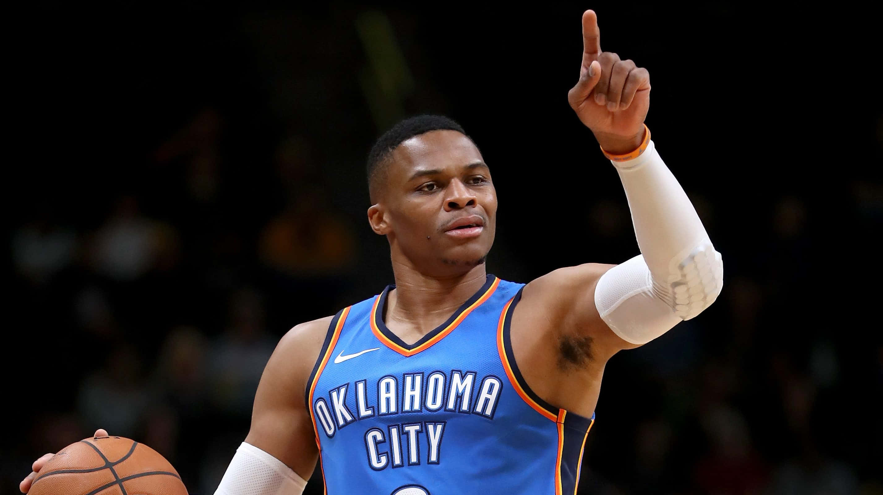 搞笑NBA指臉圖片 壁紙 — 一名身穿奧克拉荷馬城球衣的籃球運動員在場館燈光下，堅定地拿著球並向上指。