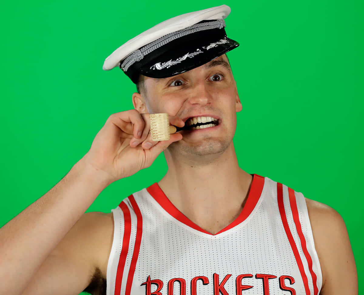 搞笑NBA水手服造型圖片 壁紙 — 一名身穿侯斯頓火箭隊球衣和船員帽的男子，在明亮的綠色屏幕前，頑皮地將牙刷放在嘴邊。