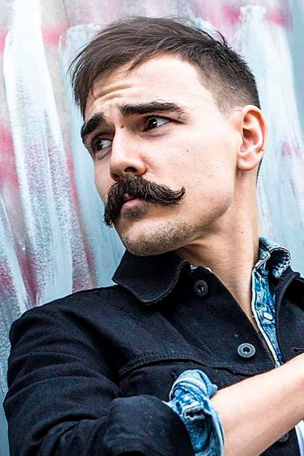 一個留著顯眼鬍鬚、身穿牛仔外套的男人，背景是塗鴉牆。