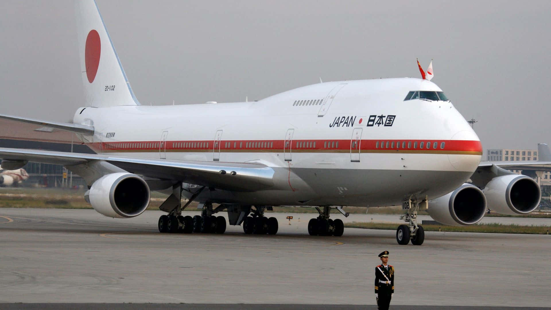 搭乘 1920x1080 分辨率巨型客機翱翔天際 壁紙 — 一架大型白色日本總統專用波音747停在機場停機坪上，附近有一名身穿制服的警衛站崗。