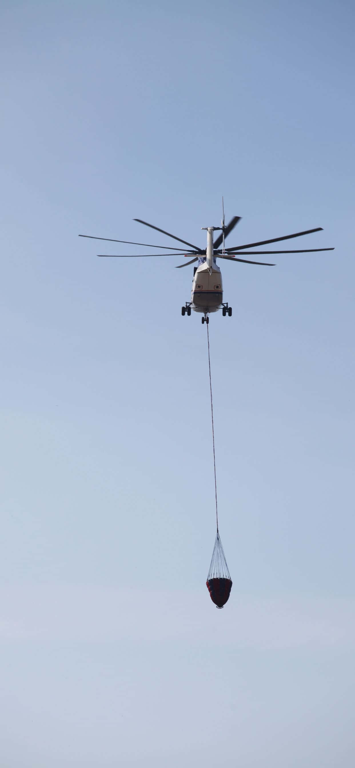 搭乘 iPhone XS Max Helicopters，享受終極空中旅程 壁紙 — 一架直升機懸掛在清澈的藍天中，用長繩放下一個紅色水桶。
