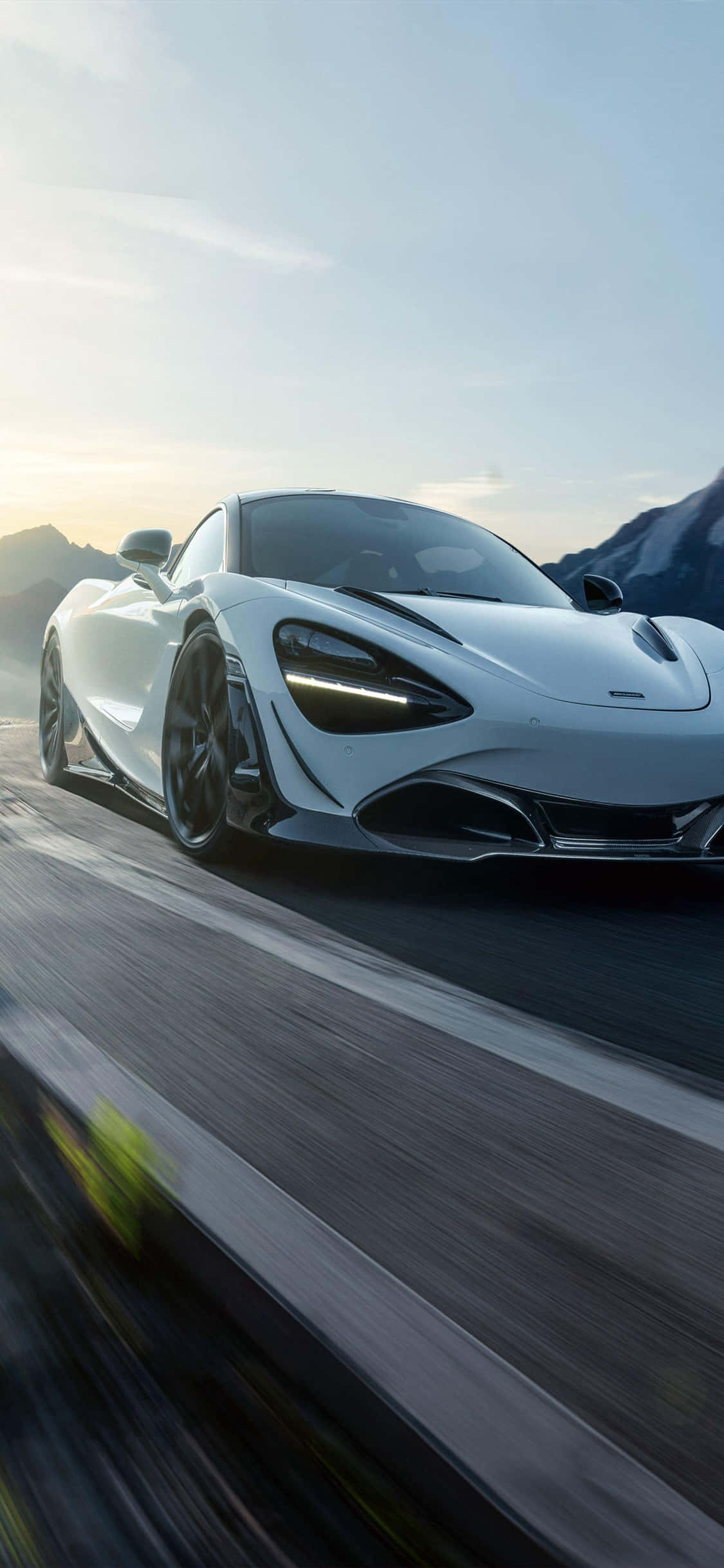 搭乘 iPhone XS 與時尚 McLaren 720S，享受優雅駕馭 壁紙 — 一輛時尚的白色跑車在日落時分沿著山路疾馳，帶有模糊的動感和遠方的山峰。