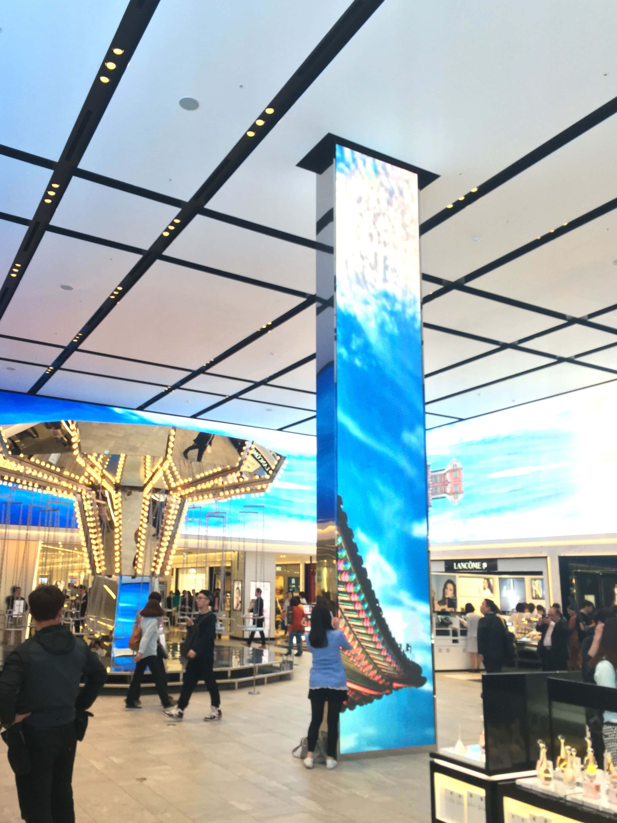 搭乘嘉年華 Vista，暢遊天堂之旅 壁紙 — 購物者在現代購物中心中庭，於高聳的數碼屏幕下漫步，屏幕顯示著鮮豔的藍天和傳統屋頂。