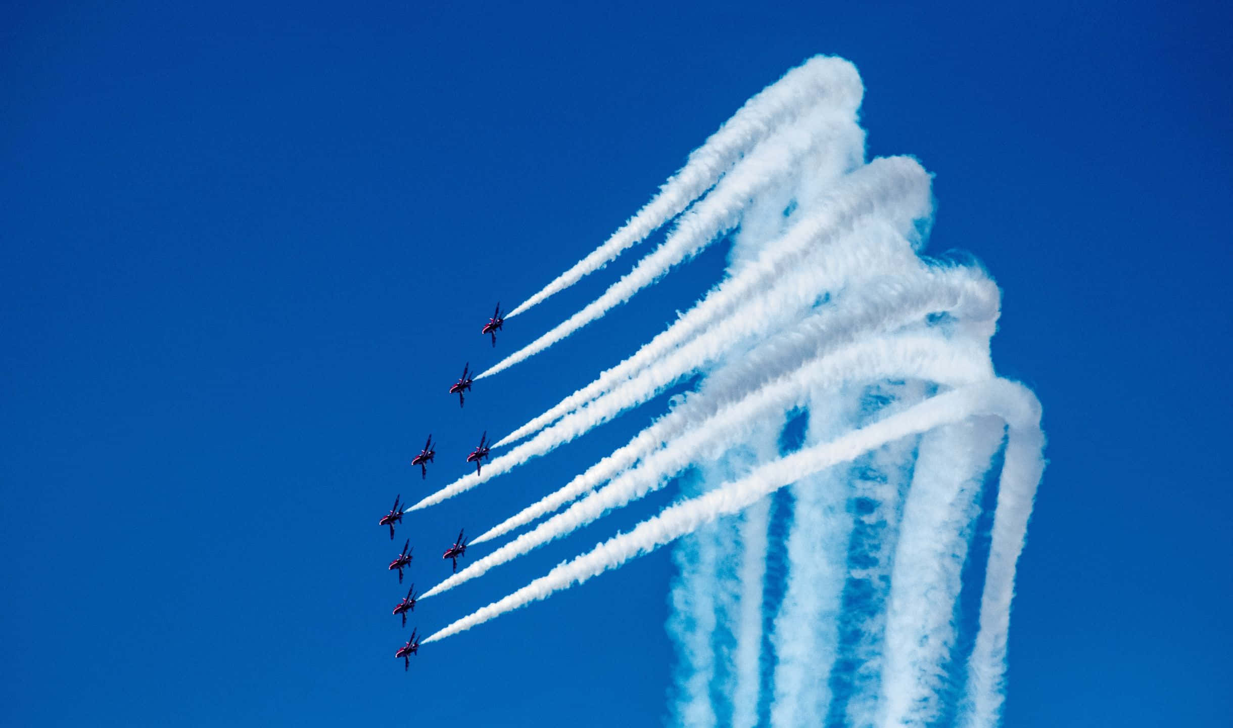 搭乘小飛機，從高空俯瞰世界 壁紙 — 九架紅色噴射機在晴朗的藍天下以緊密編隊飛行，留下濃厚的白色煙霧痕跡。