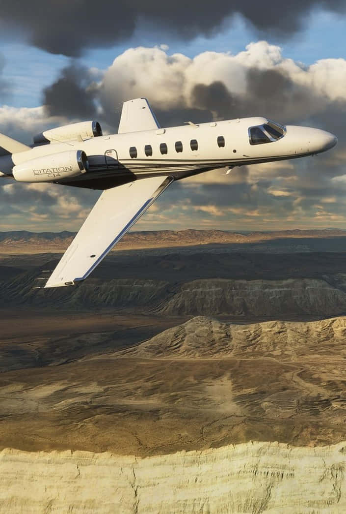 搭乘微軟飛行模擬器Android版，翱翔天際 壁紙 — 一架白色 Citation CJ4 私人飛機在戲劇性的多雲天空下，翱翔於崎嶇的沙漠峽谷之上。