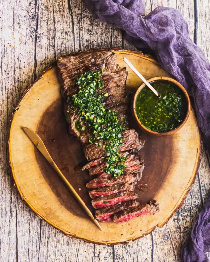 搭配新鮮奇米恰爾里醬的美味烤肉牛排 壁紙 — 切片嘅 medium-rare 牛扒，上面淋上新鮮嘅 chimichurri 醬，放喺一個 rustic 木盤上，旁邊有塊紫色布。
