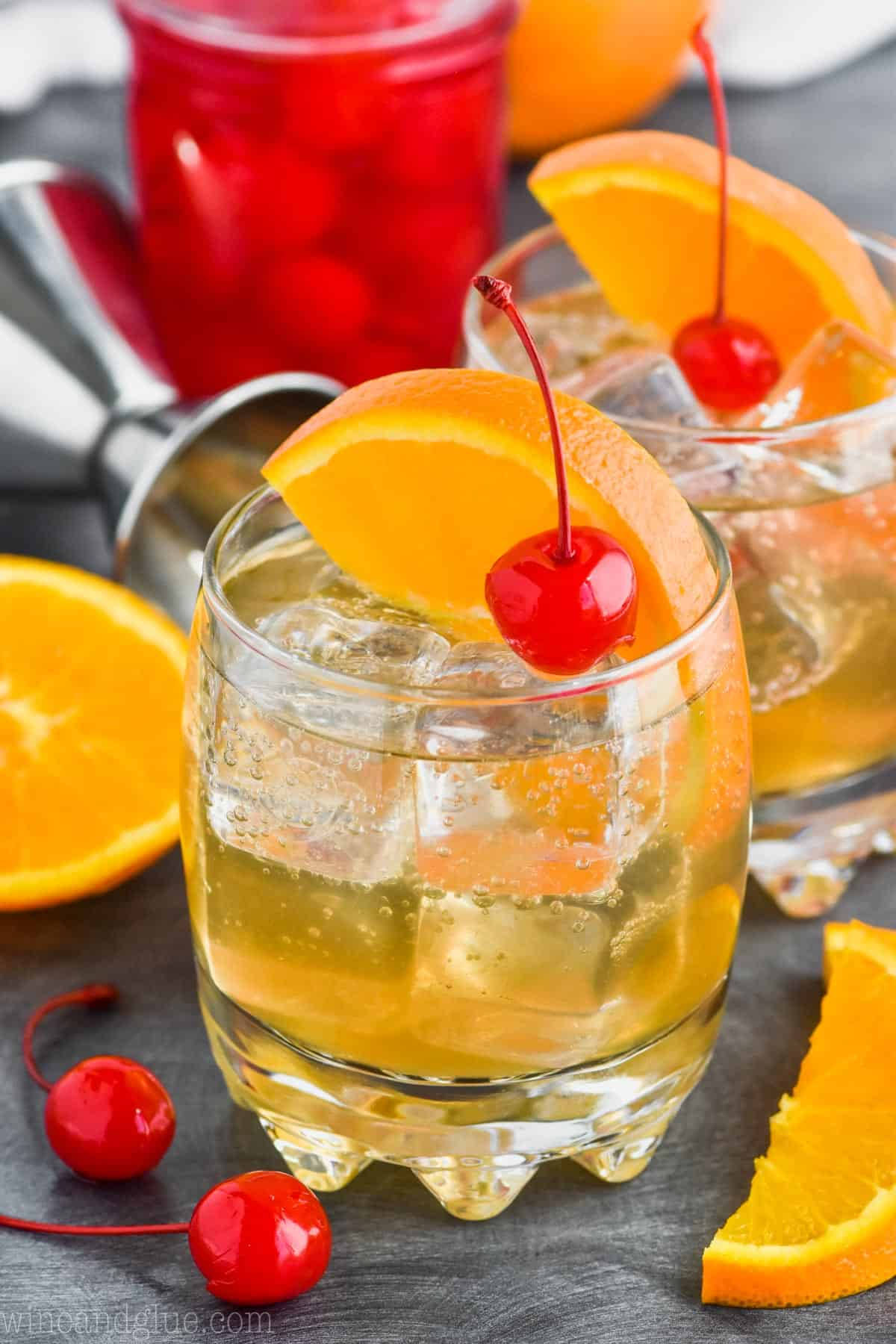 搭配水果的酸味雞尾酒 壁紙 — 兩杯琥珀色雞尾酒，配上冰塊、橙片和黑櫻桃，置於深色石板表面上。