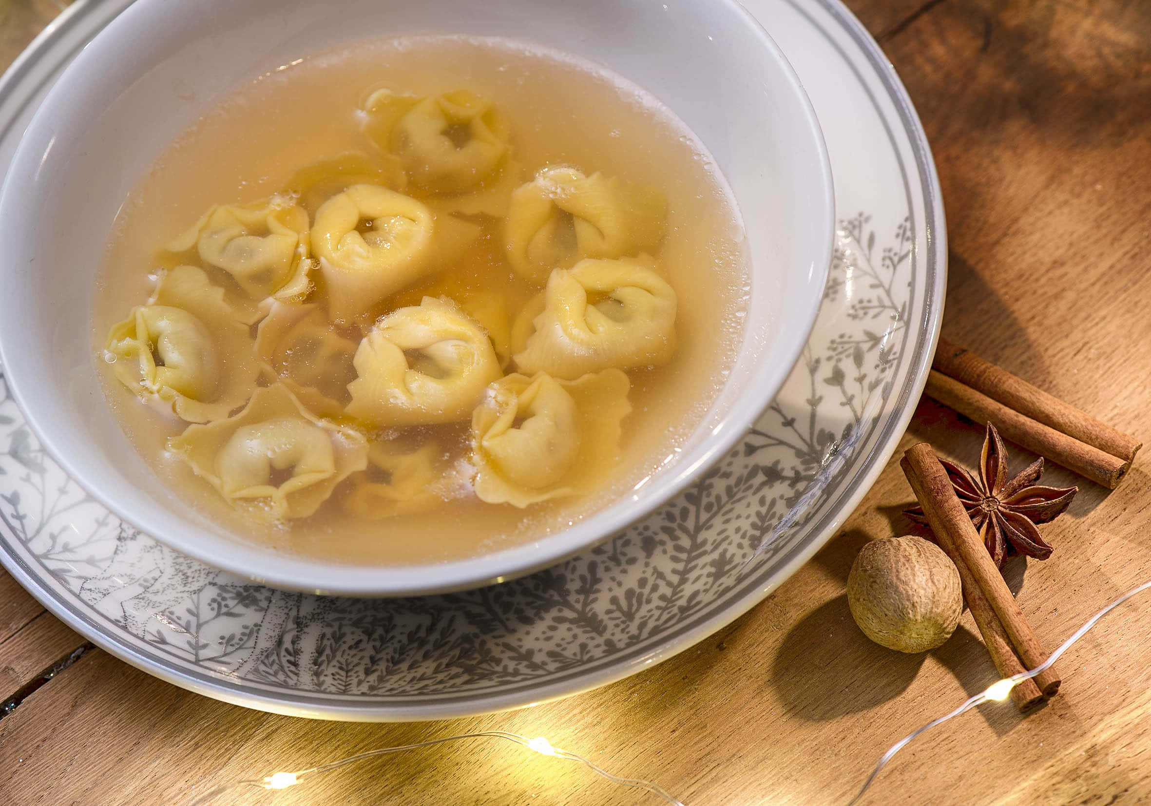 搭配肉桂捲的經典義式餃子湯 壁紙 — 一碗熱騰騰的義大利餃湯放在有圖案的碟子上，旁邊有肉桂棒、八角茴香和肉豆蔻，放在一個有仙女燈的 wooden 桌子上。