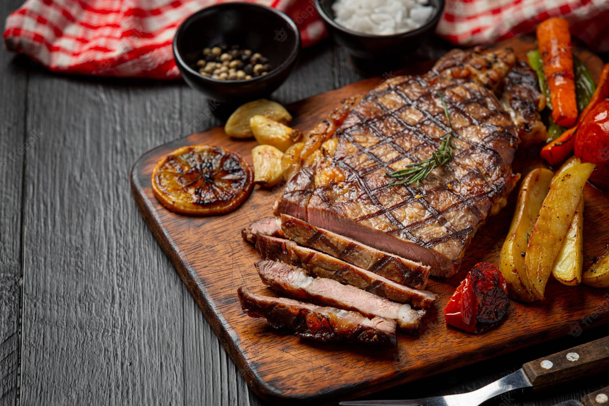 搭配馬鈴薯的烤肉Churrasco 壁紙 — 切片烤牛排帶有烤痕和迷迭香，放在木板上，旁邊有烤蔬菜和大蒜。