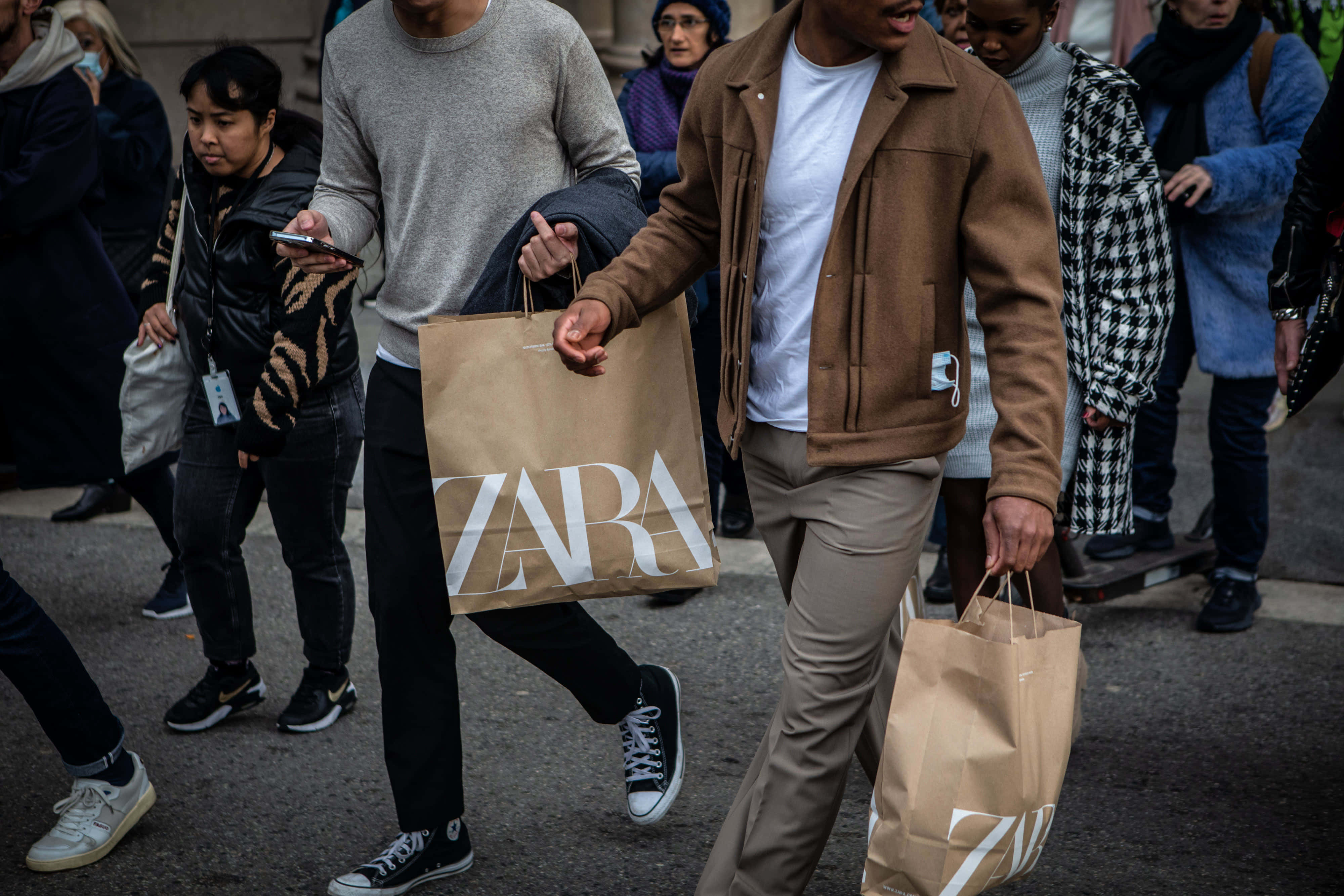 搶先體驗Zara最新潮流 壁紙 — 一名身穿棕色外套的男子，手持兩個Zara購物袋，穿過繁忙的城市街道，周圍有行人走動。