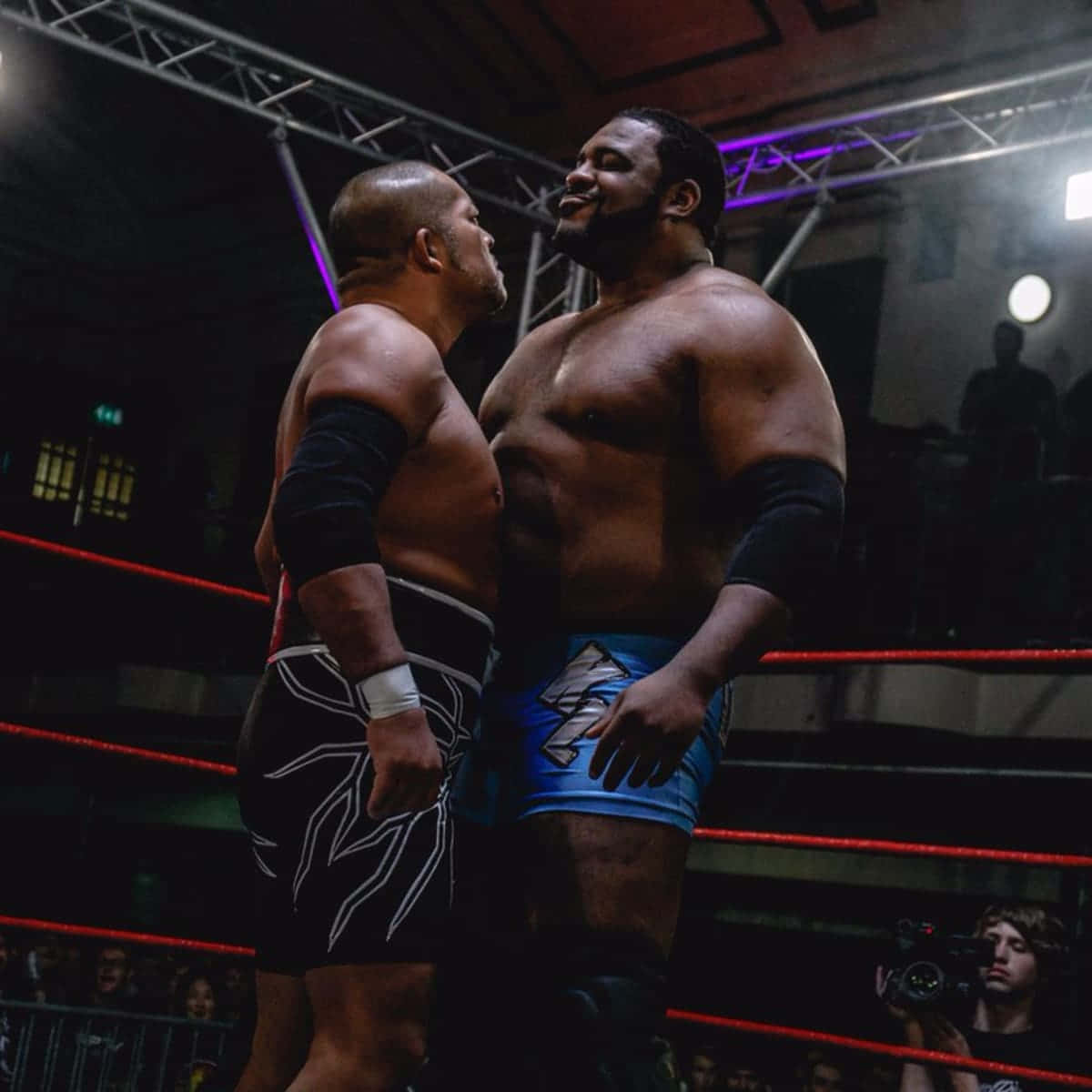 摔角戰士：Keith Lee對戰Tomohiro Ishii於Revolution Pro Wrestling賽事 壁紙 — 兩名肌肉發達的摔角手在昏暗的擂台上，在戲劇性的紫色燈光下對峙，背景有觀眾。