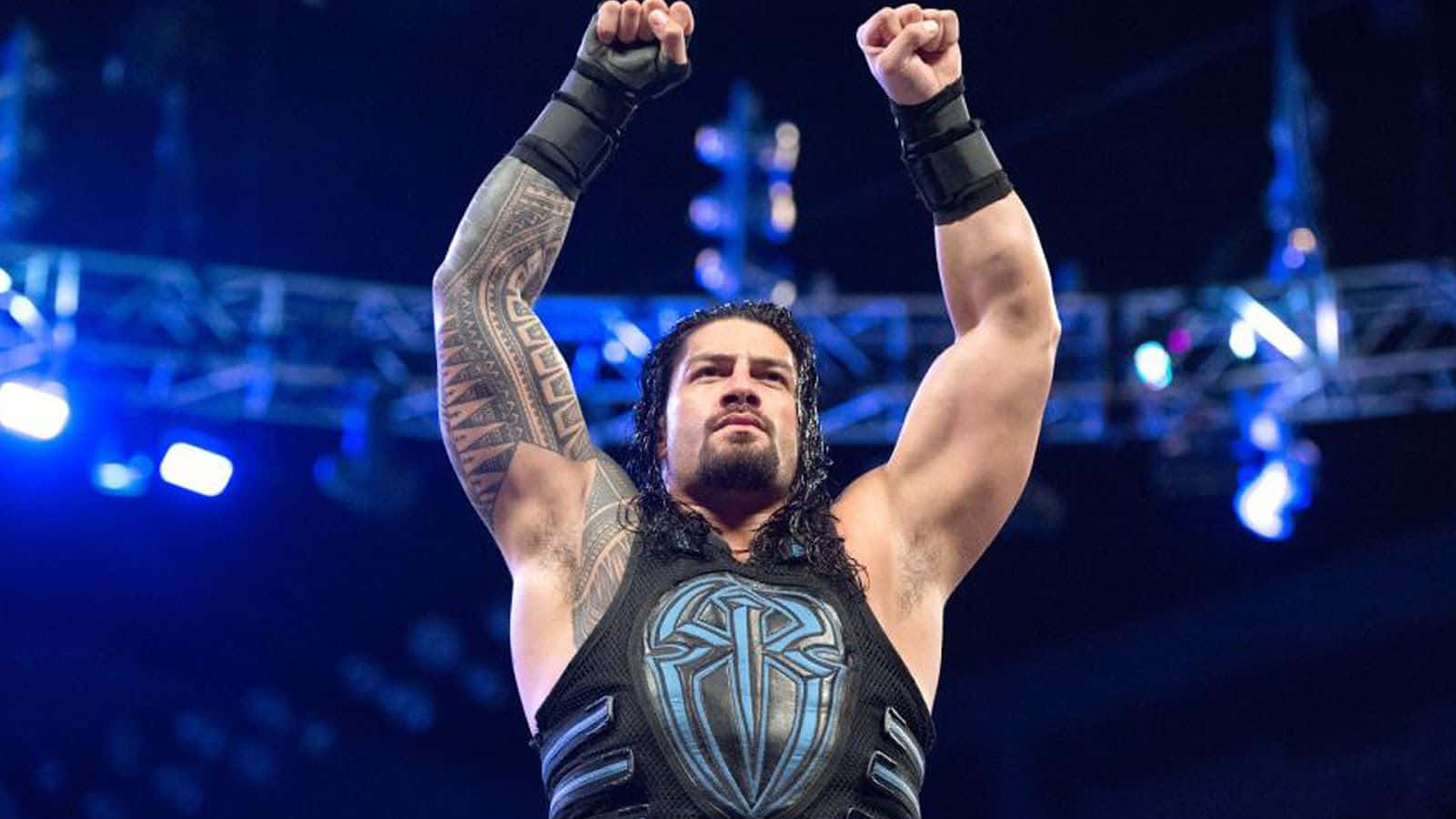 Roman Reigns 桌布 壁紙 — 摔角手 Roman Reigns 在明亮的藍色場館燈光下高舉拳頭慶祝勝利，背景是模糊的人群。