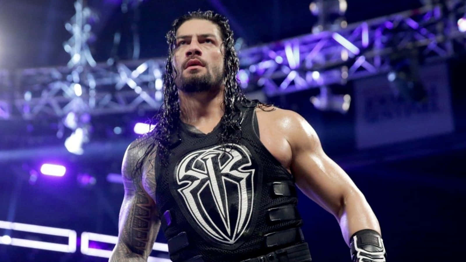 WWE 桌布 壁紙 — 摔角手 Roman Reigns 在紫色場館燈光下，身穿帶有白色標誌的黑色背心，肌肉發達，神情強烈。