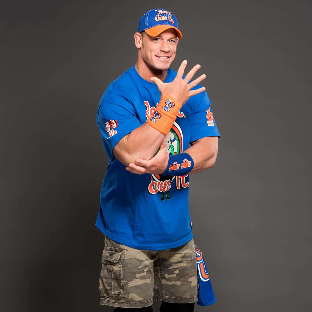 John Cena 桌布 壁紙 — 摔角手約翰·辛納在深灰色工作室背景前，穿著藍色T恤和迷彩短褲，自信地微笑。