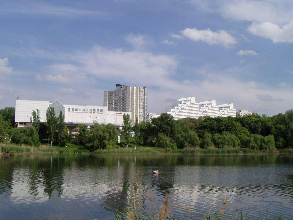 摩爾多瓦河畔建築 壁紙 — 平靜的湖面倒映著現代白色建築和茂密的綠樹，天空湛藍，白雲點點。