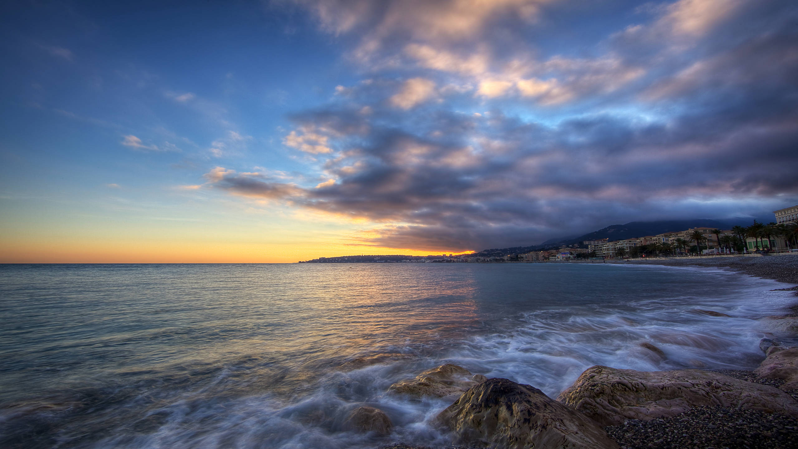摩納哥海濱夕照 壁紙 — 夕陽將天空染成金橙色和深藍色，海浪拍打著海邊小鎮旁的岩石海岸。