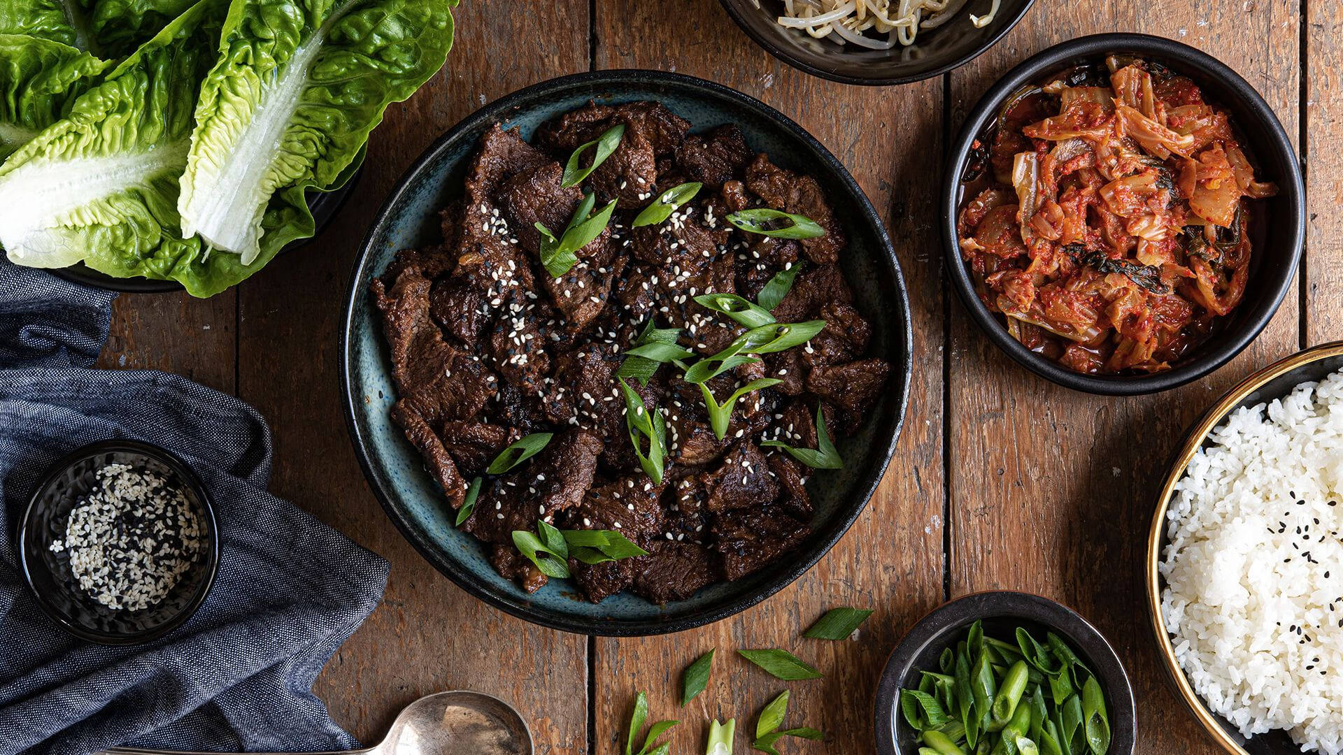 撒上芝麻的美味醬燒牛肉 壁紙 — 一張質樸的木桌上展示著韓式牛肉碗，配有泡菜飯、芝麻和新鮮生菜用於包裹。