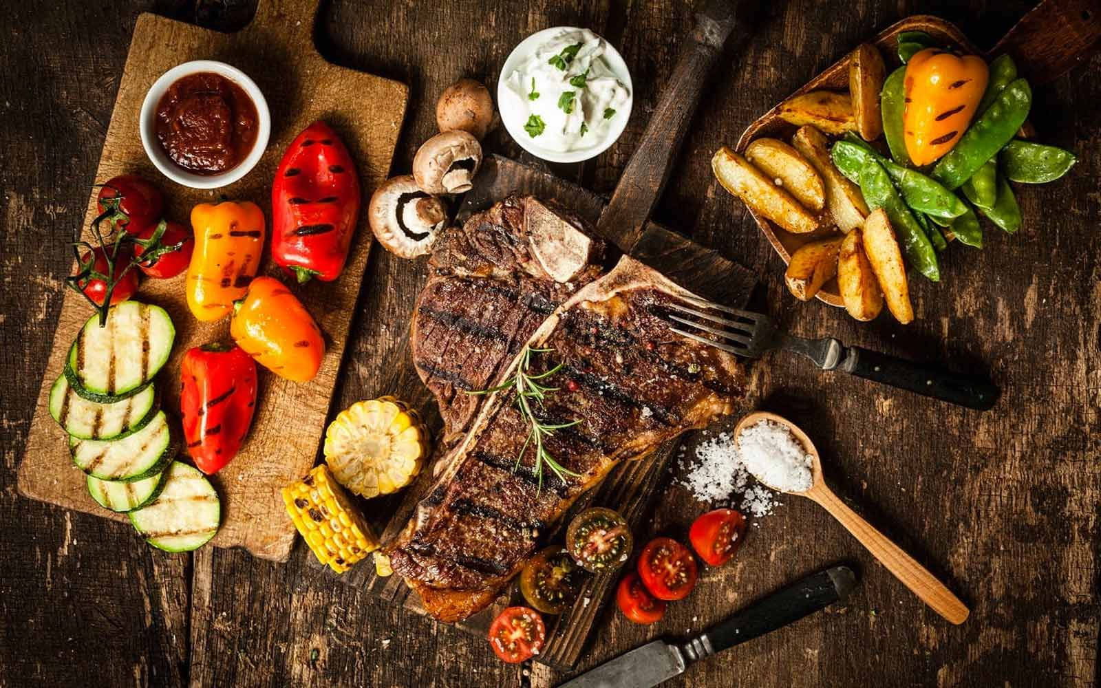 撒上香料的烤肉Churrasco 壁紙 — 多汁的烤T骨牛扒配迷迭香，放在質樸的木板上，周圍環繞著色彩繽紛的烤蔬菜和玉米。