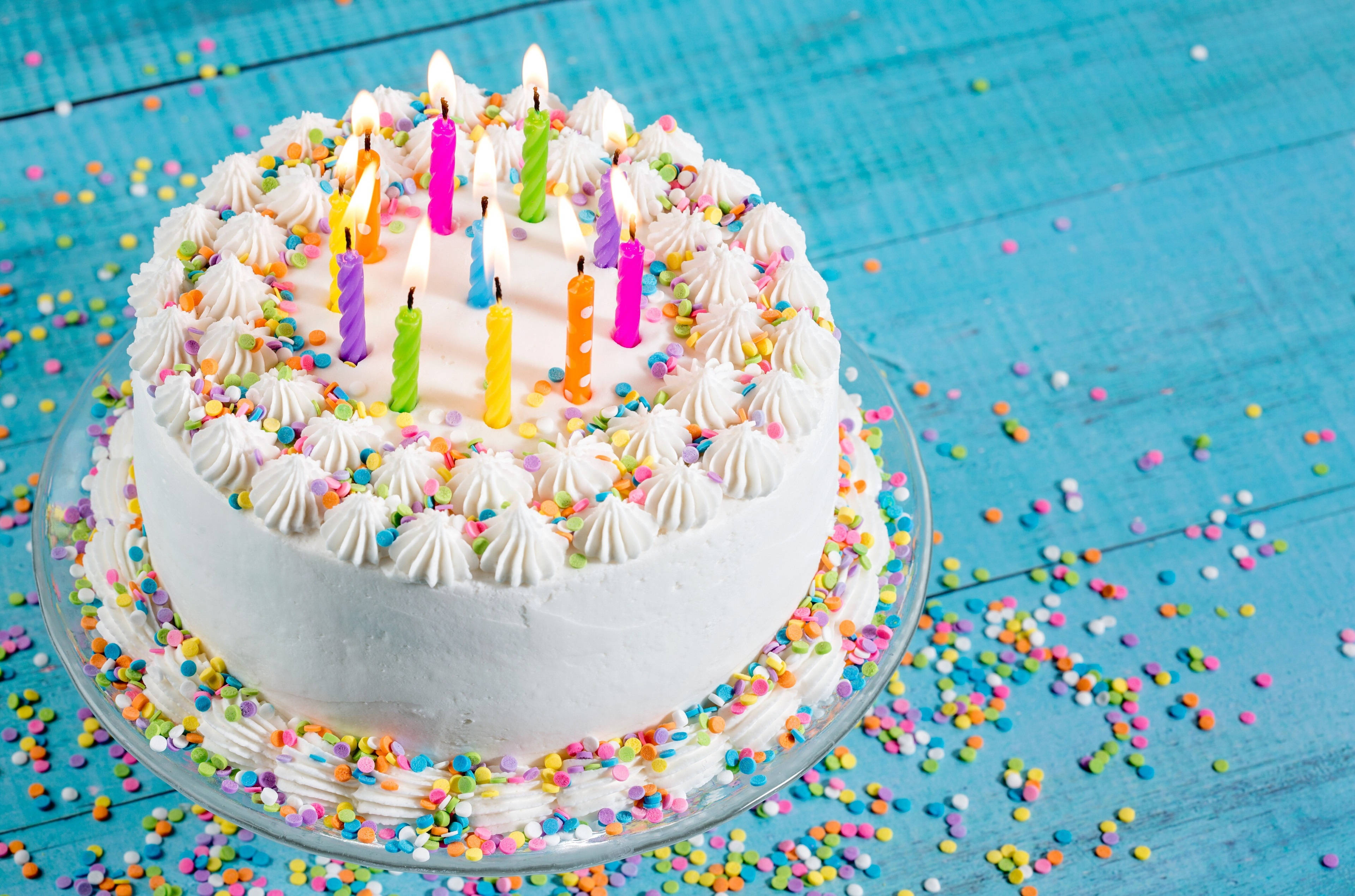 撒有糖果碎屑的生日蛋糕 壁紙 — 一個白色糖霜生日蛋糕，上面插著點燃的彩色蠟燭和彩虹糖針，放在藍色木桌上。