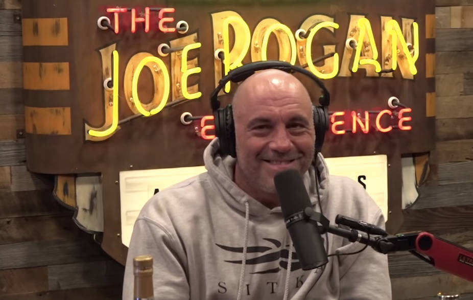 Joe Rogan 喺podcast錄音室入面微笑，身後有霓虹燈牌。