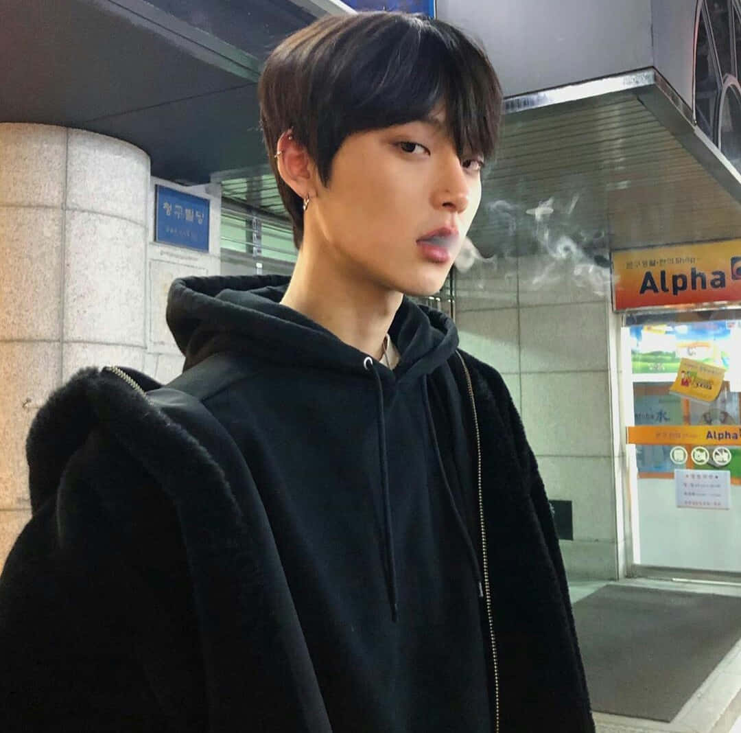 擁抱你的時尚風格 壁紙 — 一名穿著黑色連帽衫的年輕男子在燈光昏暗的都市車站中呼出煙霧，車站有韓文標誌。