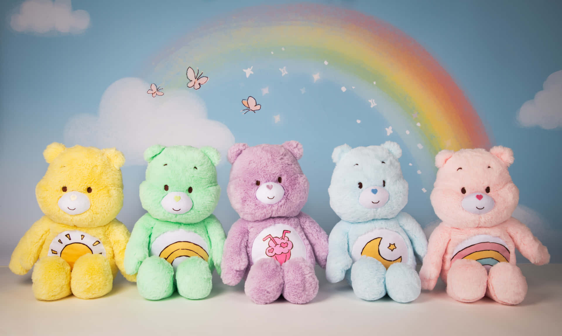 五隻Care Bears毛絨玩具在彩虹下排成一排，伴有蝴蝶。