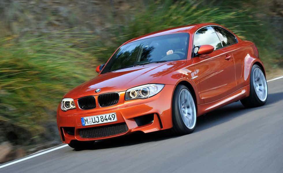 橙色BMW M1雙門跑車在彎曲的道路上疾馳，兩旁是綠色的山丘。