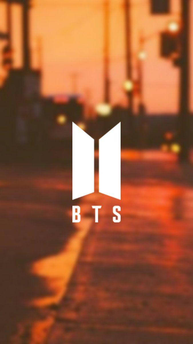 擁抱柔和橙色美學的BTS氛圍 壁紙 — 白色BTS標誌居中於黃昏時分模糊的橙色街景，帶有濕漉漉的人行道倒影。
