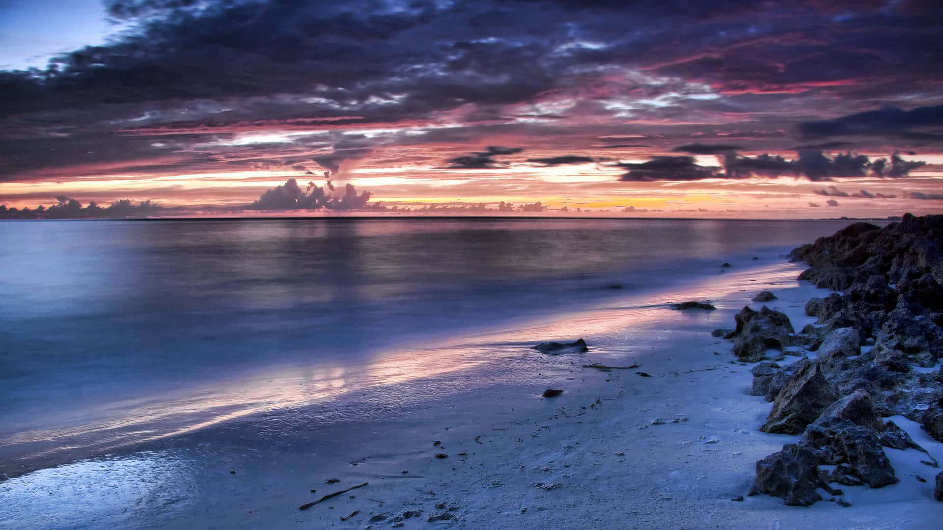 擁抱海灘上的月光 壁紙 — 日落時分的寧靜海灘，鮮豔的紫色和橙色雲彩倒映在岩石海岸旁的平靜水面上。