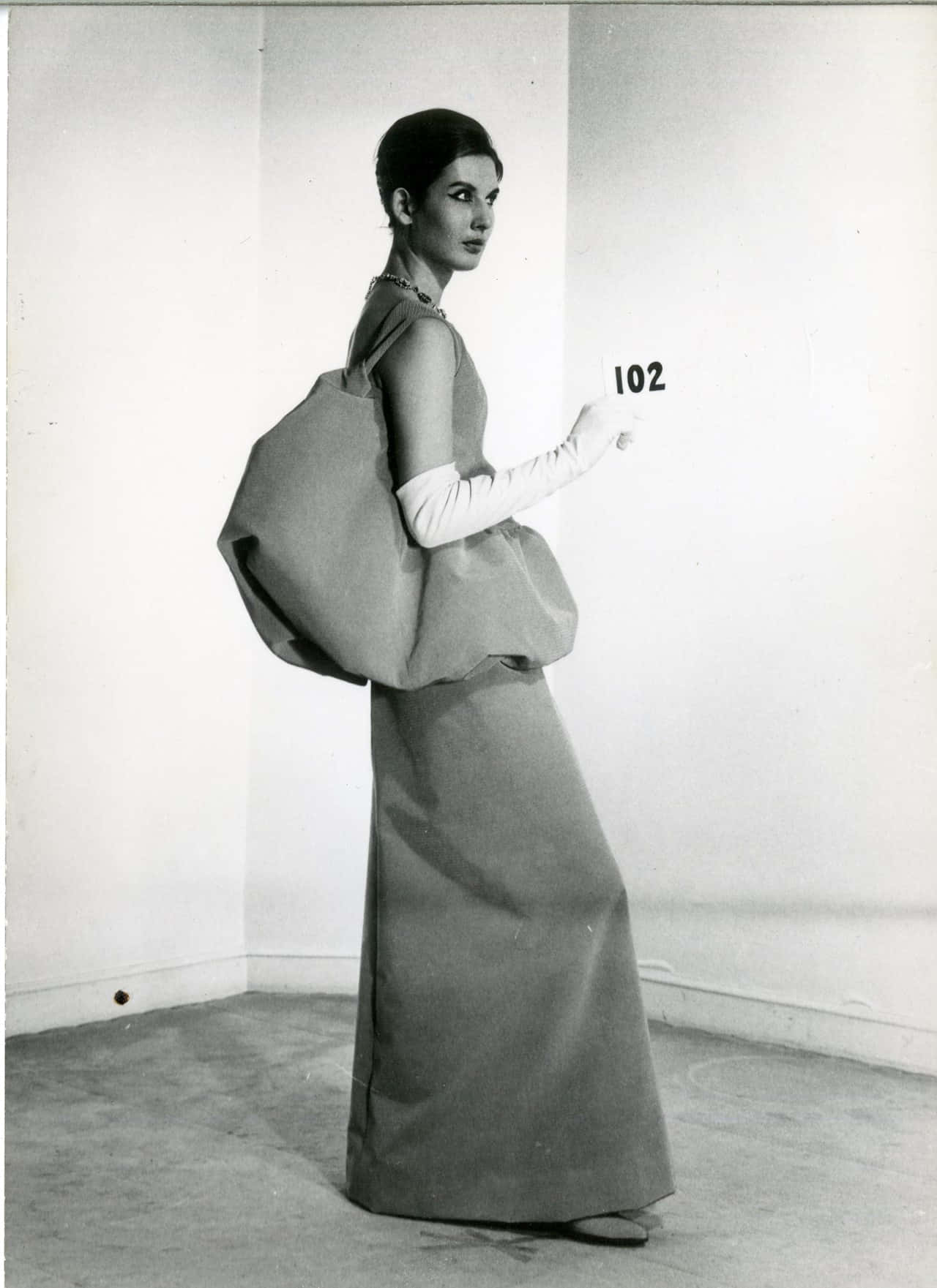 擁抱現代美學，選擇Balenciaga 壁紙 — 一位優雅女性身穿1960年代蓬鬆連衣裙和白色手套，站在極簡工作室角落，手持號碼牌。