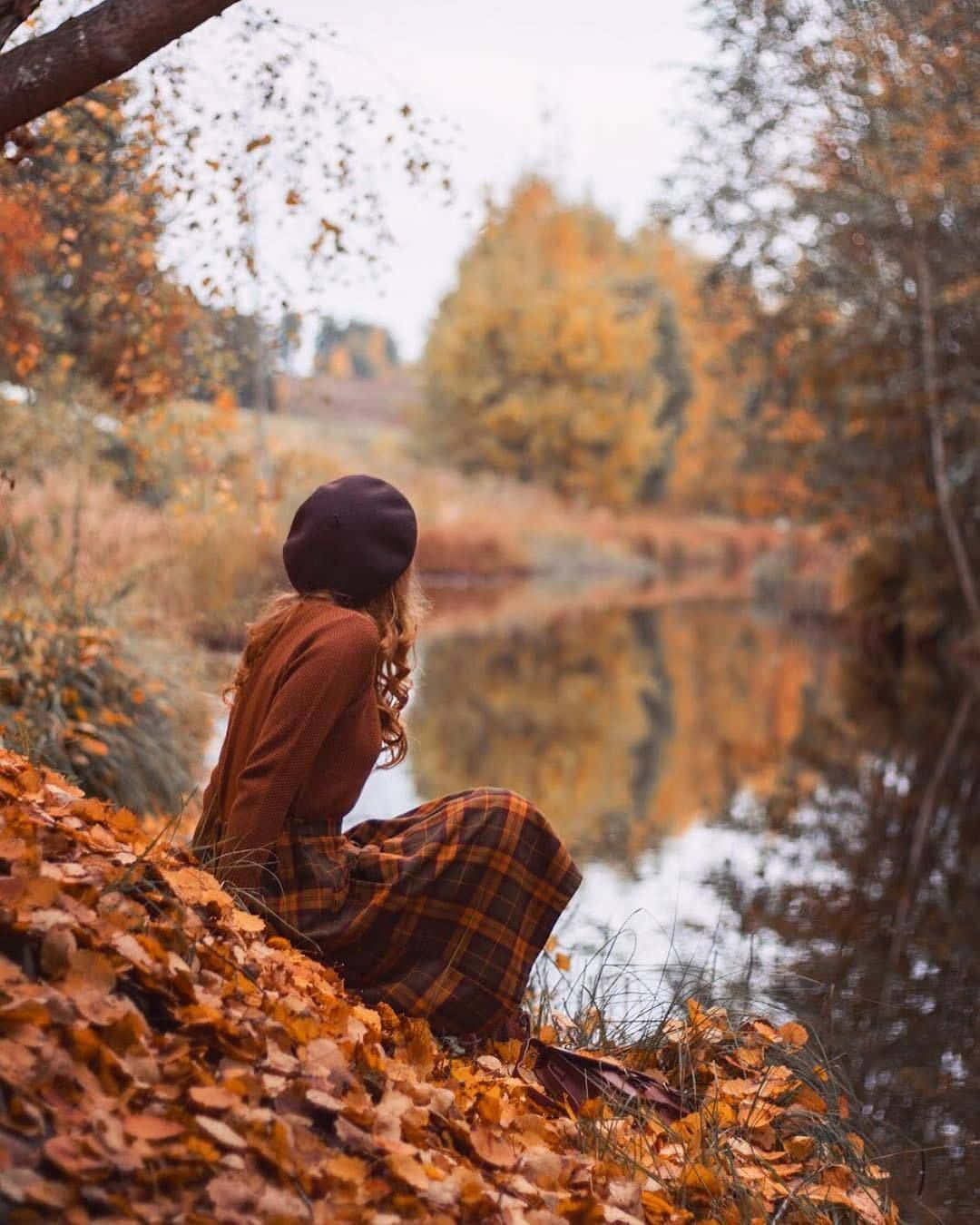 擁抱秋日葉片變幻的絕美色彩 壁紙 — 一位戴貝雷帽、穿格子裙的女子坐在落葉堆上，凝視著倒映秋樹的平靜河流。
