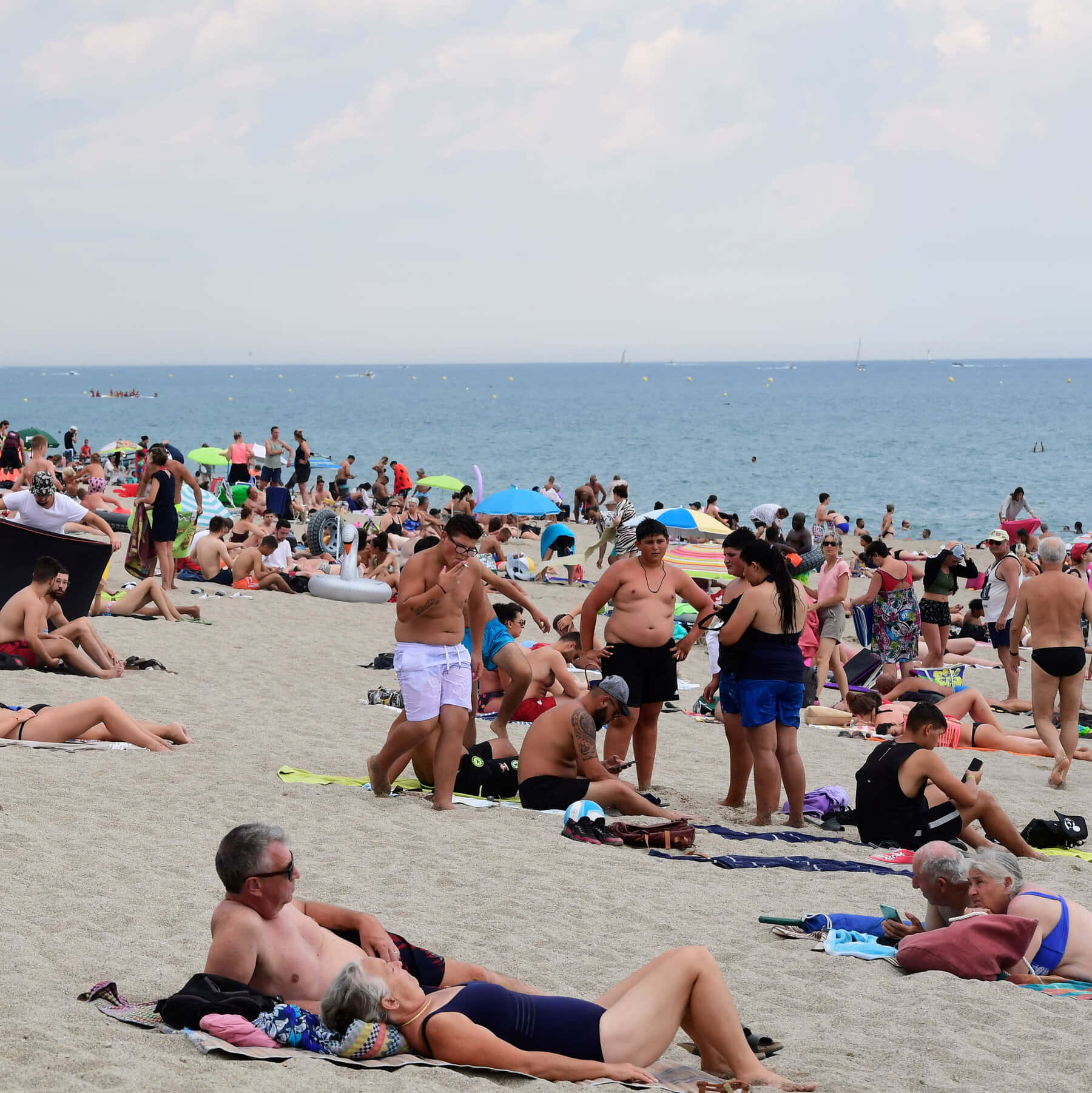 French Beach 桌布 壁紙 — 擁擠的海灘場景，日光浴者在明亮的天空下在沙灘上放鬆，海洋延伸至地平線。