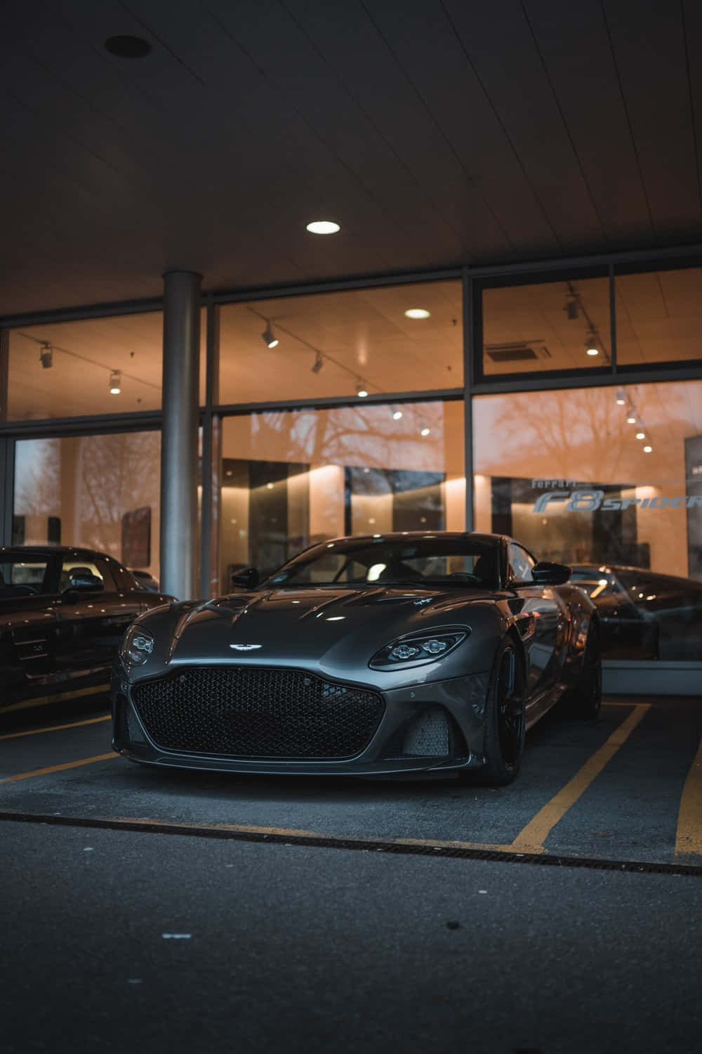擁有一輛阿斯頓·馬丁，體驗奢華生活 壁紙 — 一輛時尚的啞光灰色 Aston Martin Vantage 在夜間停放在現代玻璃天篷下，溫暖的車內燈光反射在其拋光表面上。