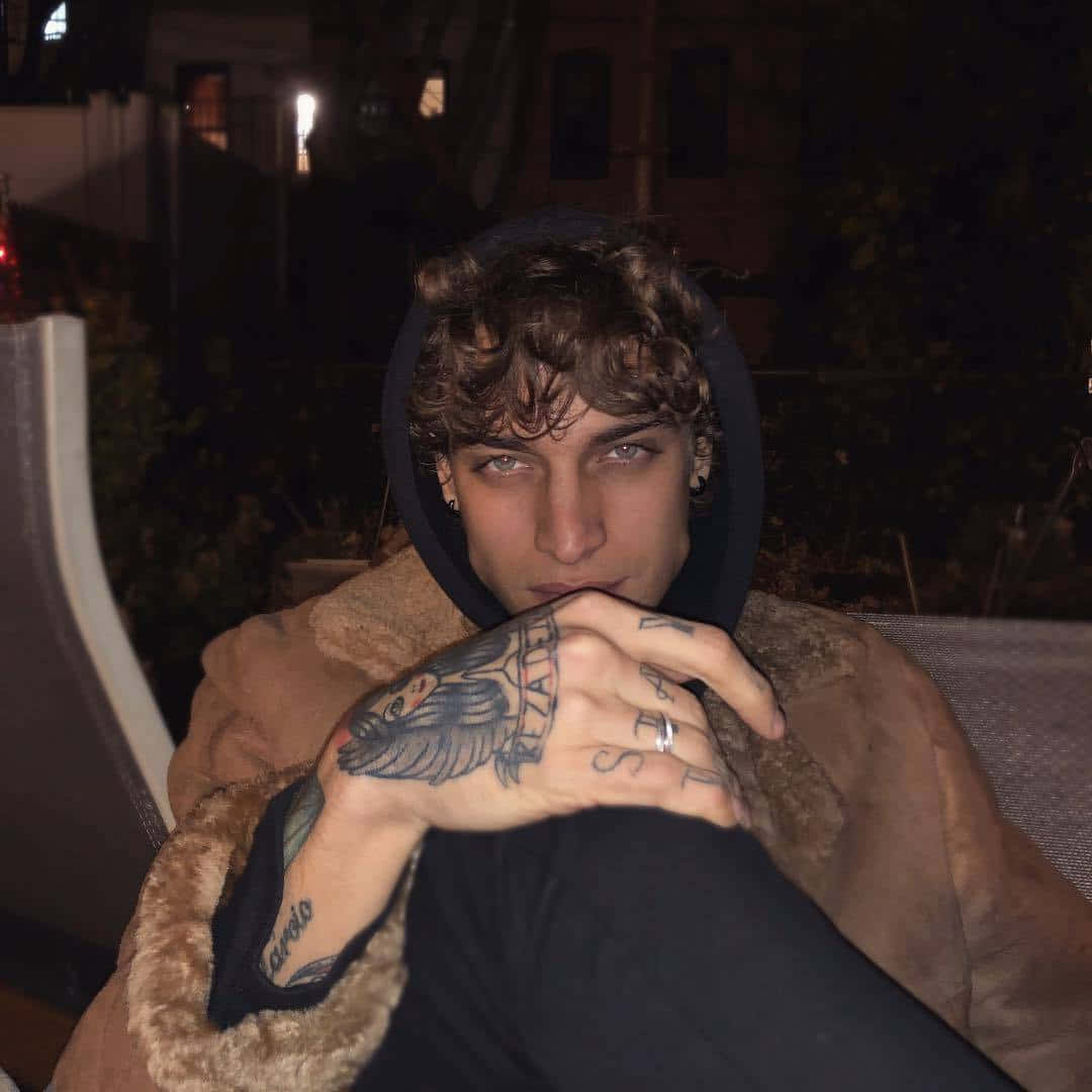 擁有大膽精緻紋身的年輕男子 壁紙 — 一個捲髮且有紋身的年輕男子，在夜晚將有紋身的手放在連帽衫下的臉旁休息。