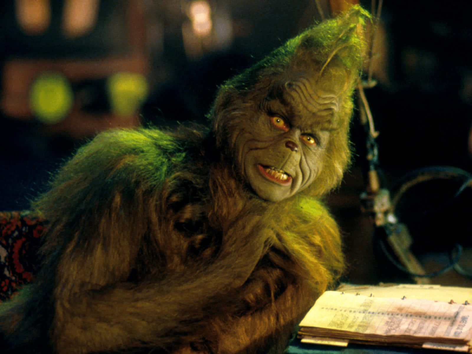 The Grinch 桌布 壁紙 — 擁有發光黃色眼睛和綠色毛髮的鬼靈精，在昏暗的房間裡威脅性地坐在雜亂的書桌前。